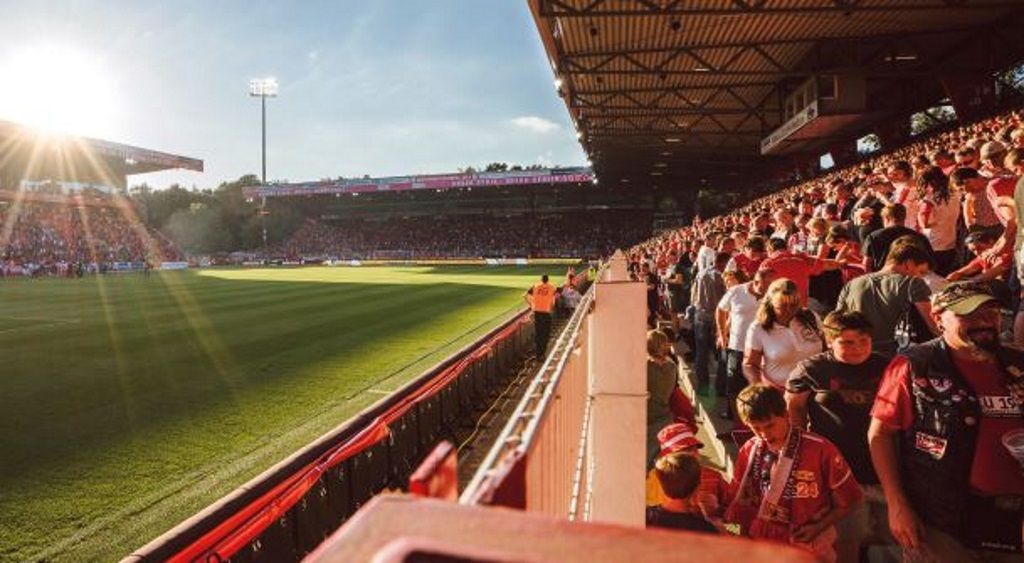 Ein vollbesetztes Stadion bei Sonnenuntergang, mit jubelnden Fans und einem grünen Spielfeld im Vordergrund.