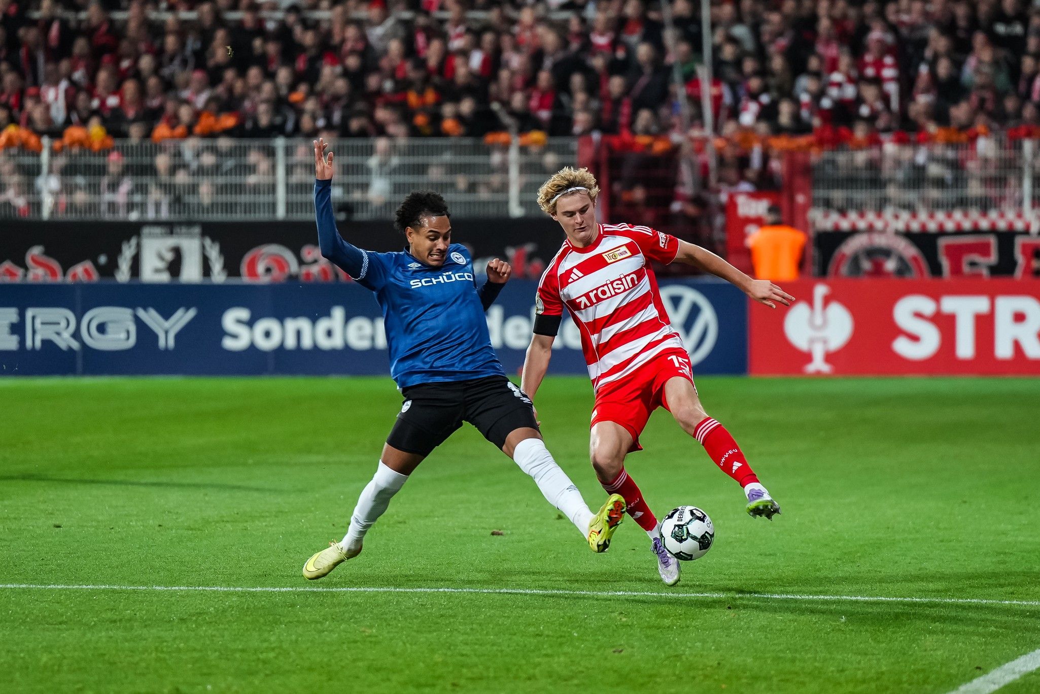 A football player in blue and a player in red are fighting for the ball on a stadium field, surrounded by spectators.