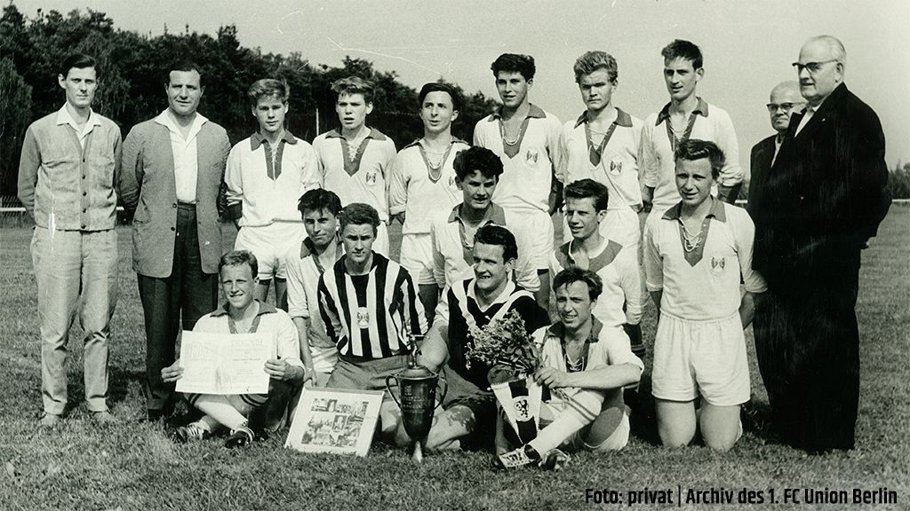 Schwarz-weißes Foto einer Fußballmannschaft mit Pokalen, umgeben von Spielern und einem Trainer auf einem Sportplatz.