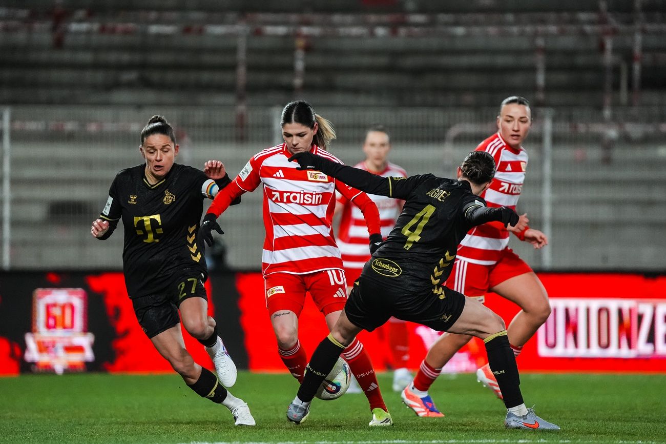 Spielszene im Frauenfußball mit zwei Spielerinnen in schwarzer Trikots und einer Spielerin in rotem Trikot, die den Ball kontrolliert.