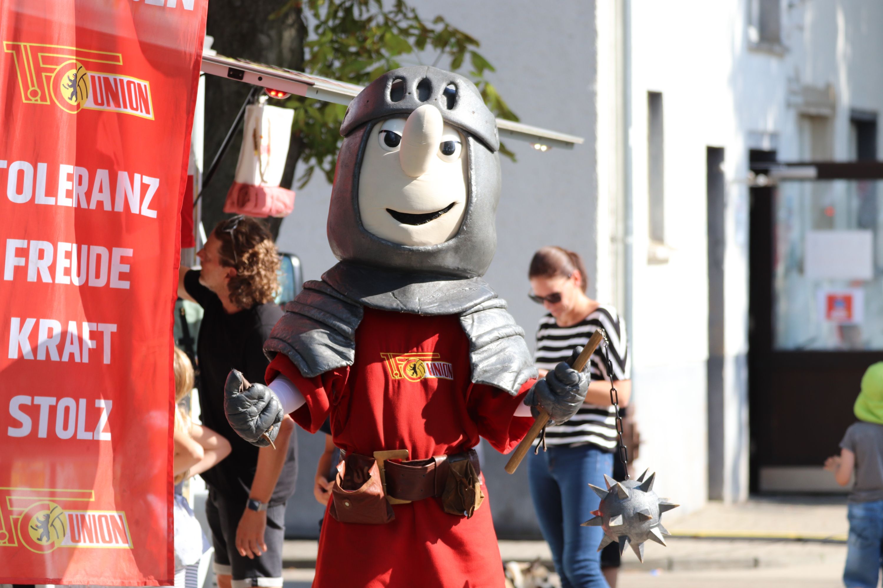 Ein Maskottchen in Ritterrüstung mit einem großen Kopf steht an einem Stand, umgeben von Menschen und einer roten Fahne.