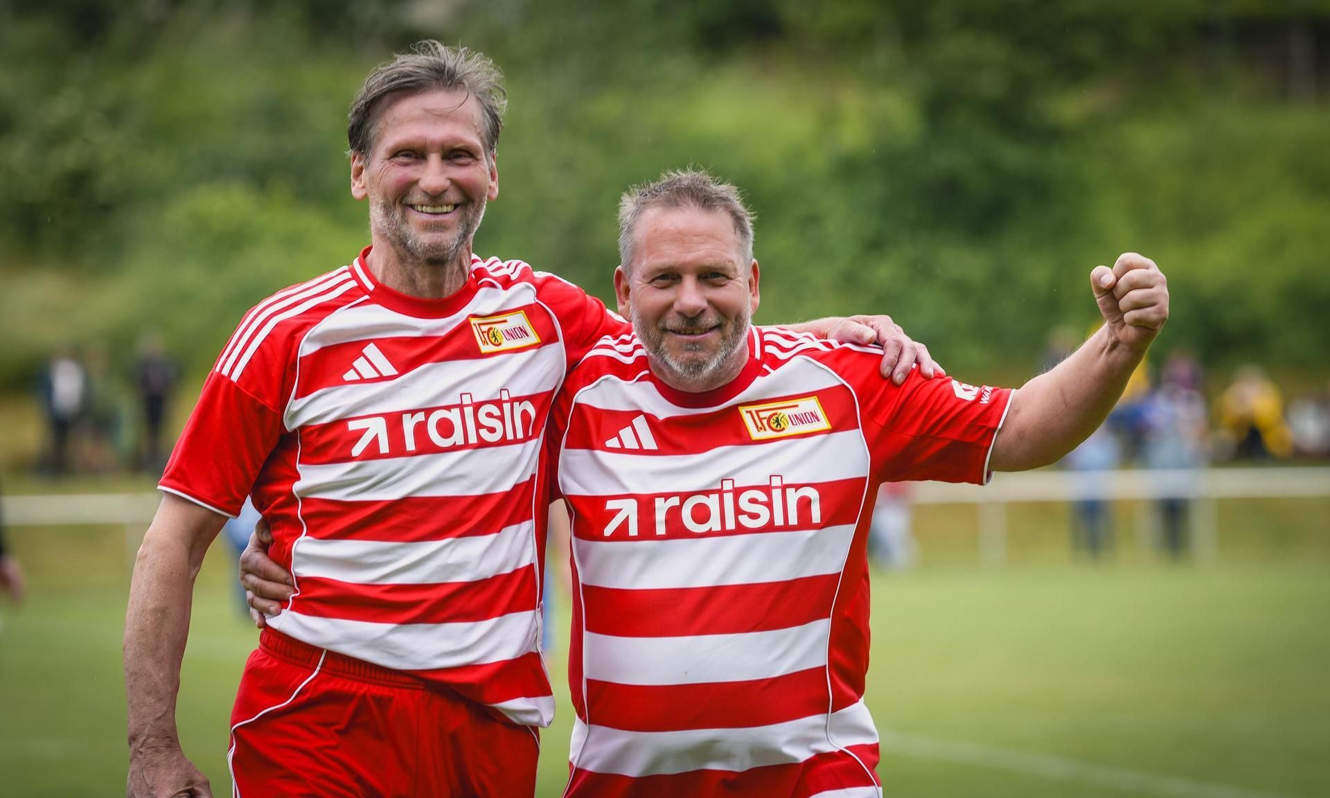 Zwei lächelnde Fußballspieler in roten gestreiften Trikots, die sich umarmen und den Sieg feiern.