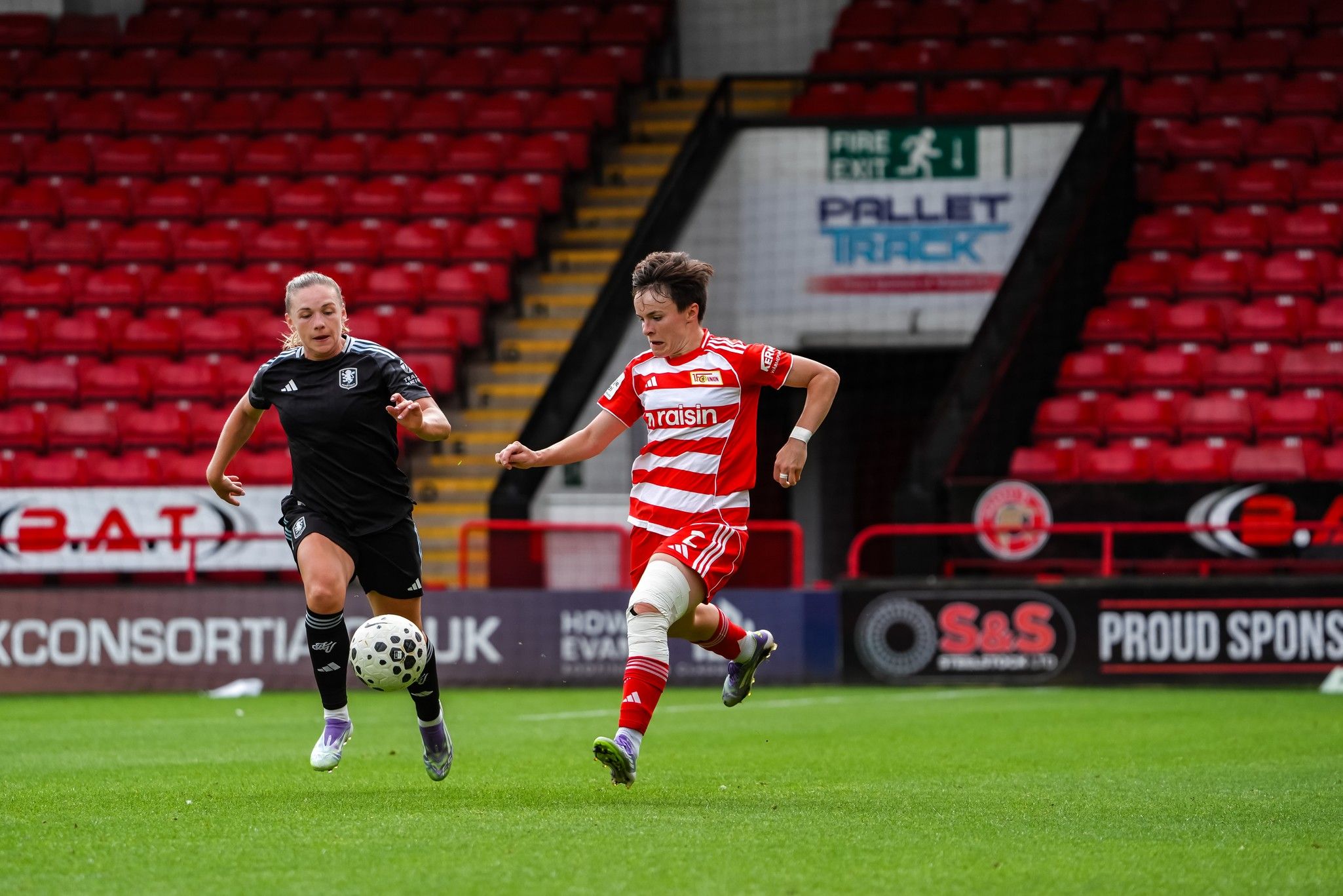 Zwei Fußballspielerinnen in Action, eine trägt schwarz und die andere rot-weiß gestreifte Trikots, auf einem Rasenplatz.