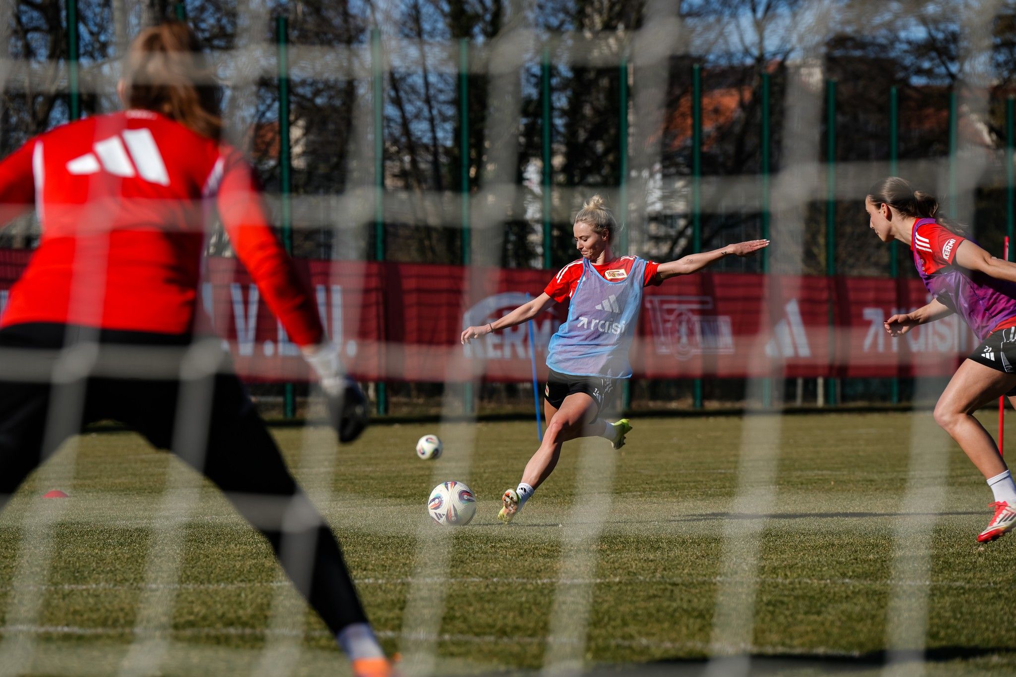 Spielerin tritt den Ball während des Fußballtrainings, mit zwei weiteren Spielerinnen im Hintergrund und dem Tor im Fokus.