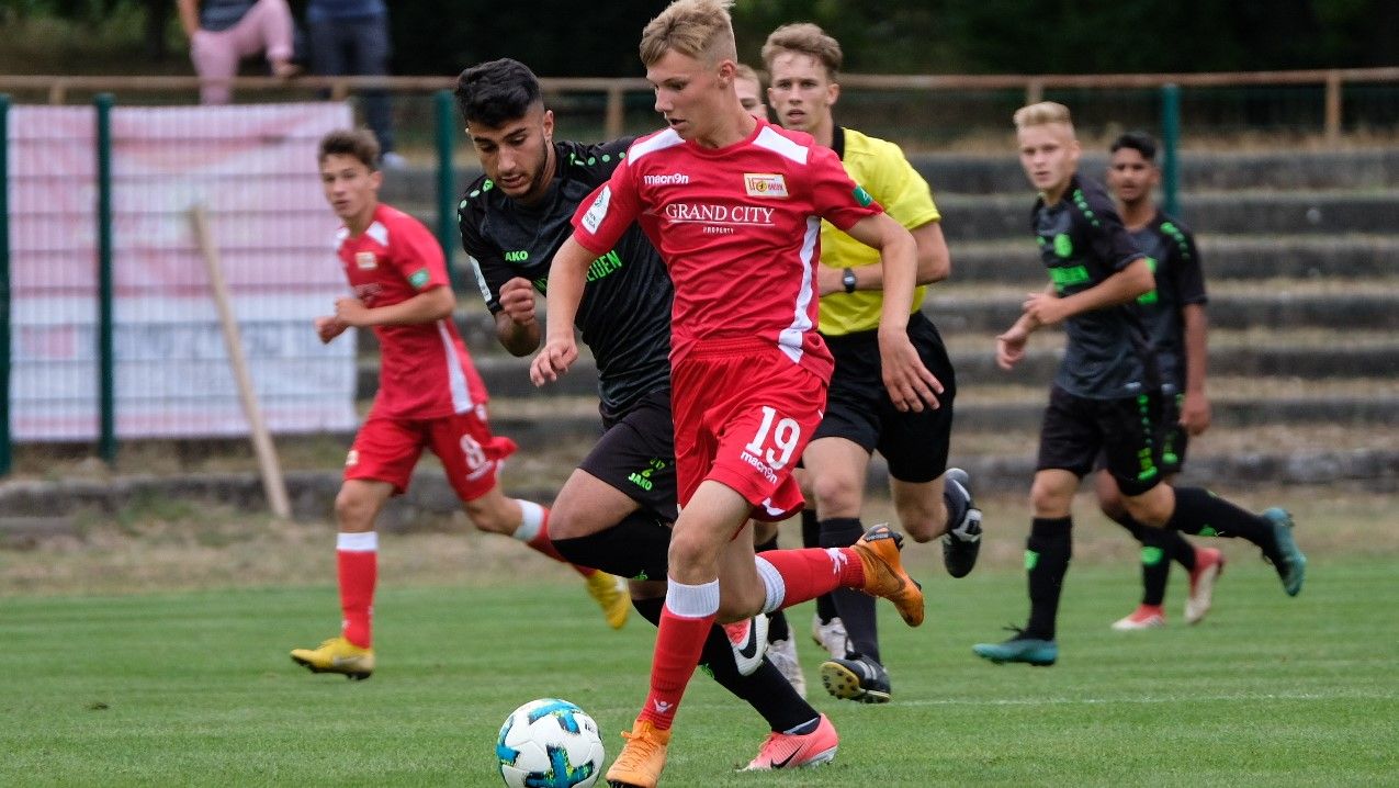 Ein junger Fußballspieler trägt ein rotes Trikot. Er dribbelt den Ball. Andere Spieler laufen hinter ihm her. Es ist eine Szene auf einem Rasenplatz.