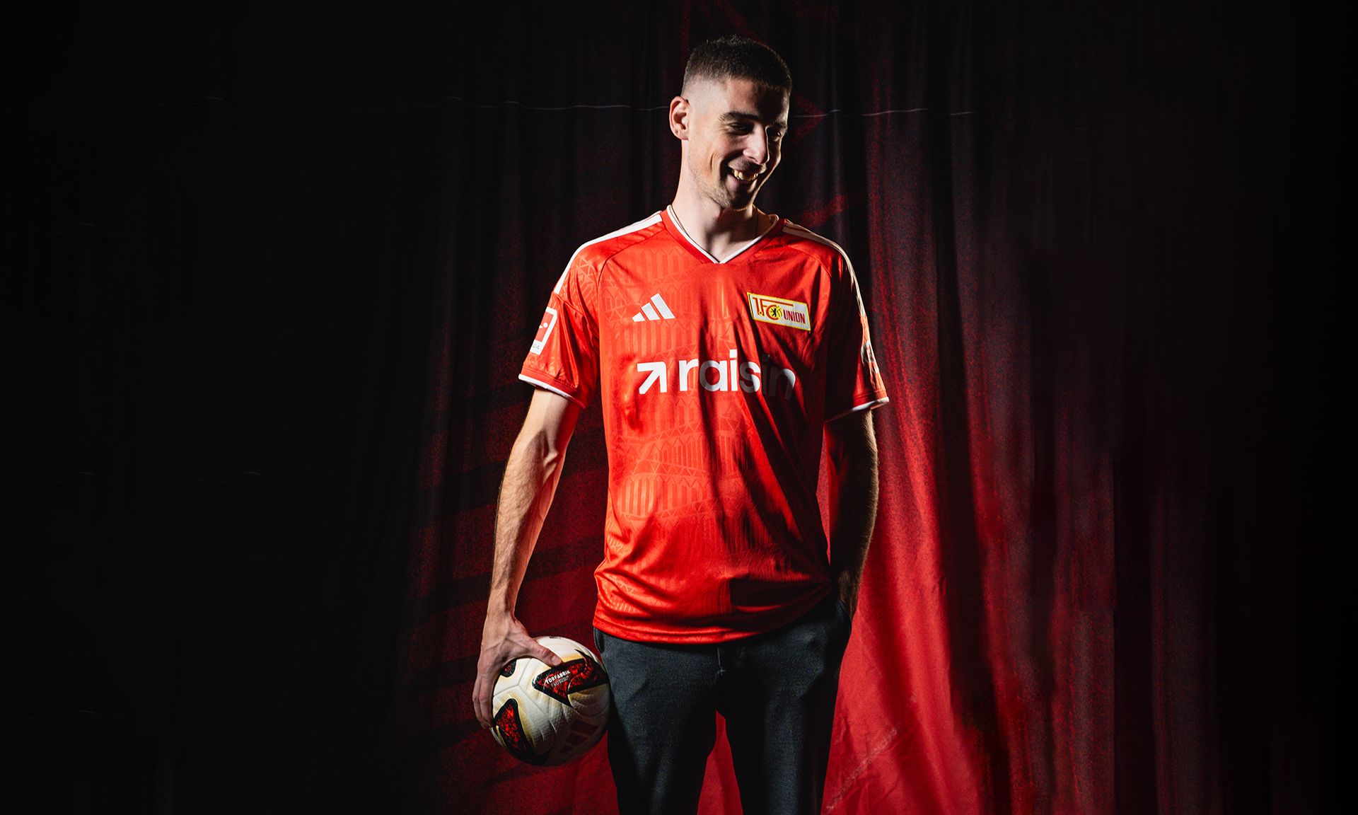 Ein Mann trägt ein rotes Fußballtrikot, hält einen Ball in der Hand und steht vor einem schwarzen Hintergrund mit roten Akzenten.