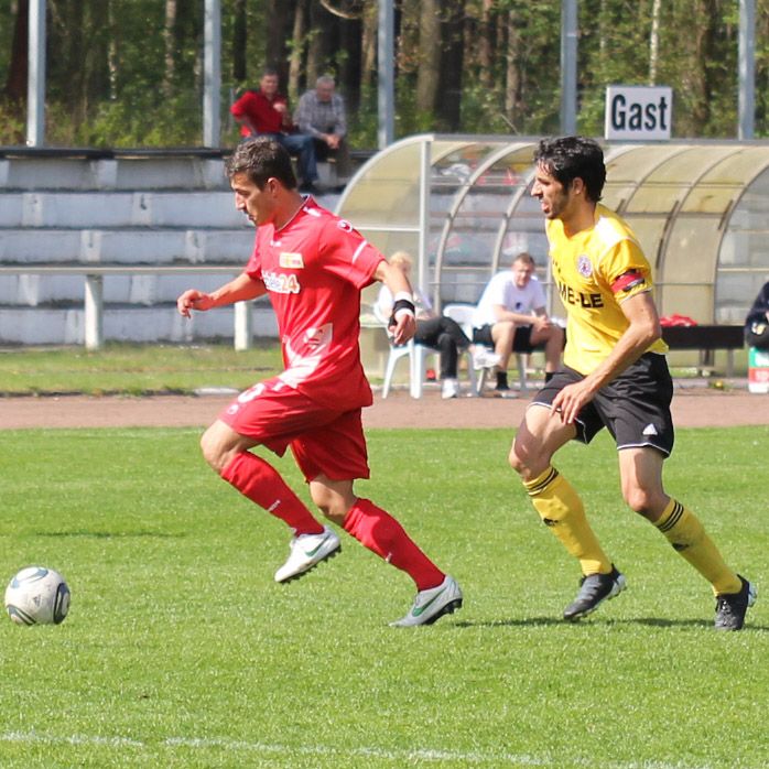 Zwei Fußballspieler in roten und gelben Trikots laufen auf einem Rasenplatz, während ein Zuschauerbereich im Hintergrund sichtbar ist.