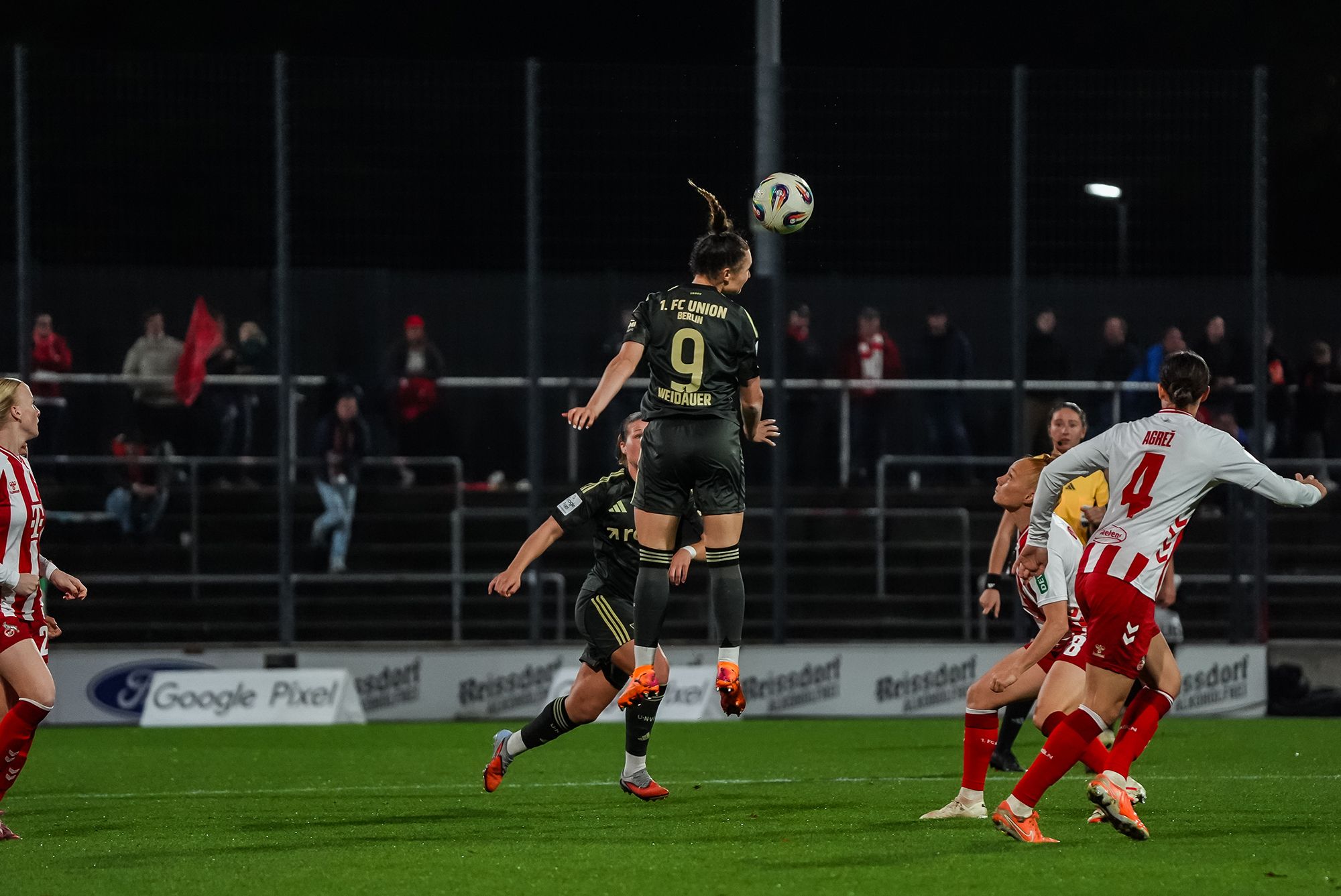 Eine Spielerin springt in einem Fußballspiel hoch, während sie einen Ball köpft, umgeben von Gegnerinnen auf dem Spielfeld.