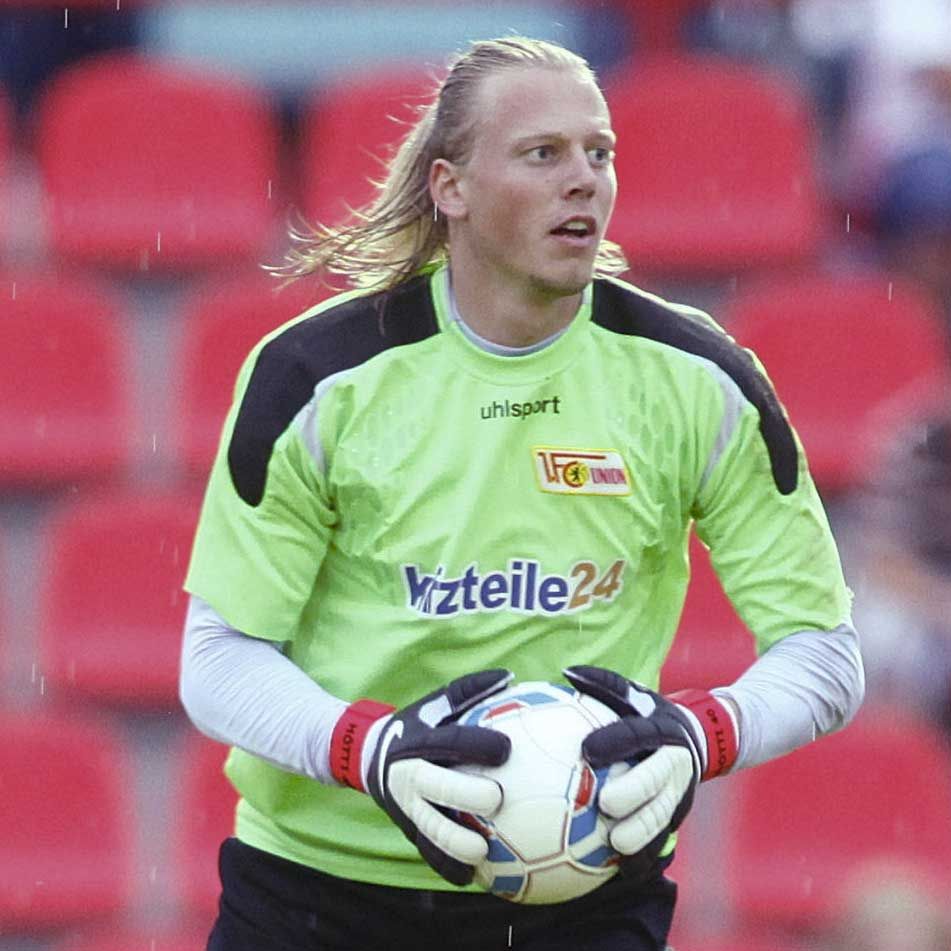 Torhüter in grünem Trikot hält einen Fußball, mit langen Haaren und ernster Miene, im Stadion unter Regen.