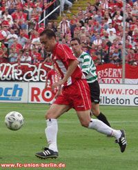 Ein Fußballspieler in roter Kleidung dribbelt den Ball, während ein Gegner in Schwarz-Weiß ihn bewacht; im Hintergrund jubelt die Menge.