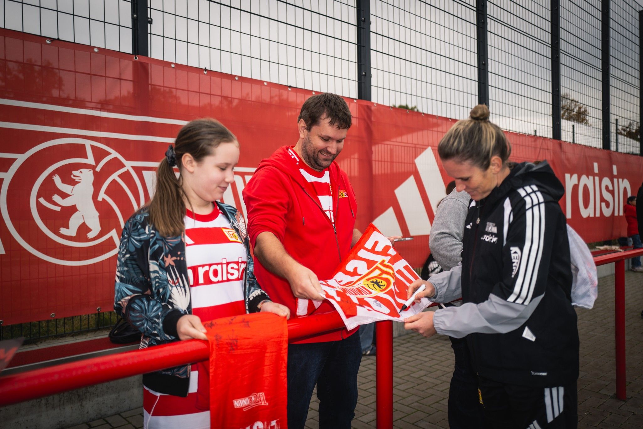 Zwei Fans und eine Person in Sportkleidung tauschen ein Fan-Poster vor einer Stadionwand aus.