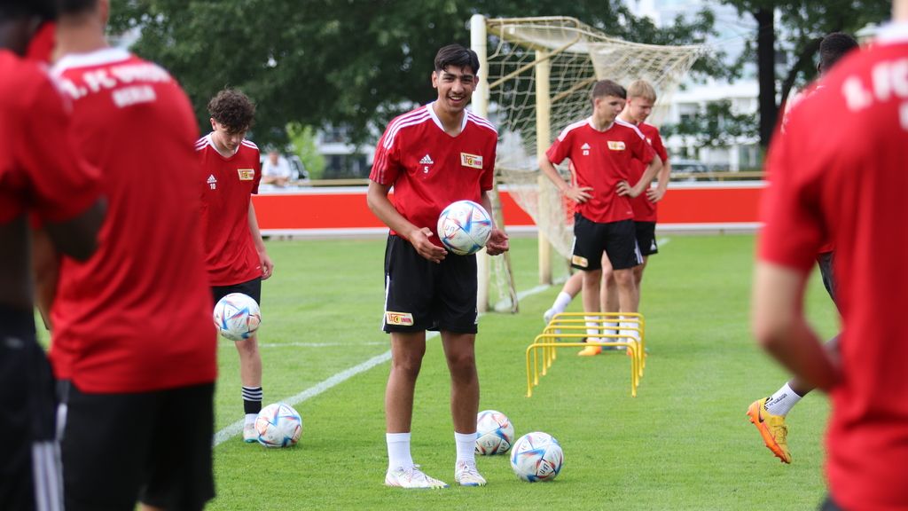 Junge Fußballspieler in roten Trikots trainieren auf einem grünen Platz mit Bällen und Hütchen im Hintergrund.