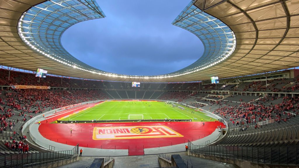 Leeres Stadion mit roten Sitzen und einem grünen Fußballfeld, beleuchtet unter einem bewölkten Himmel.