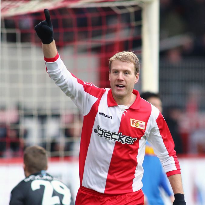 Ein Fußballspieler mit kurzem blondem Haar und rot-weißem Trikot zeigt mit dem Finger nach oben, während er jubelt.