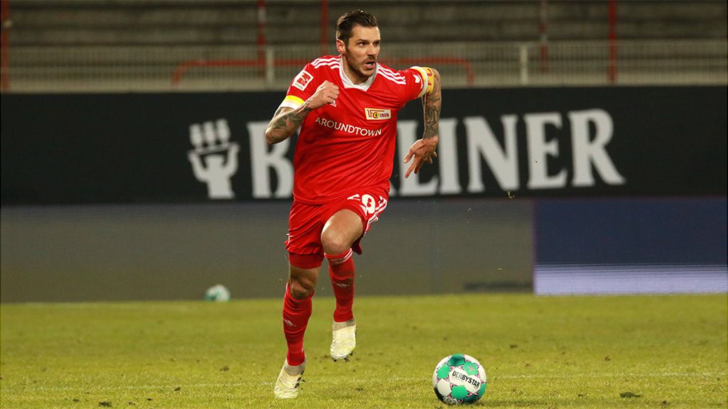 Ein Fußballspieler trägt ein rotes Trikot. Er läuft schnell über das Feld. Im Hintergrund steht eine Werbetafel.