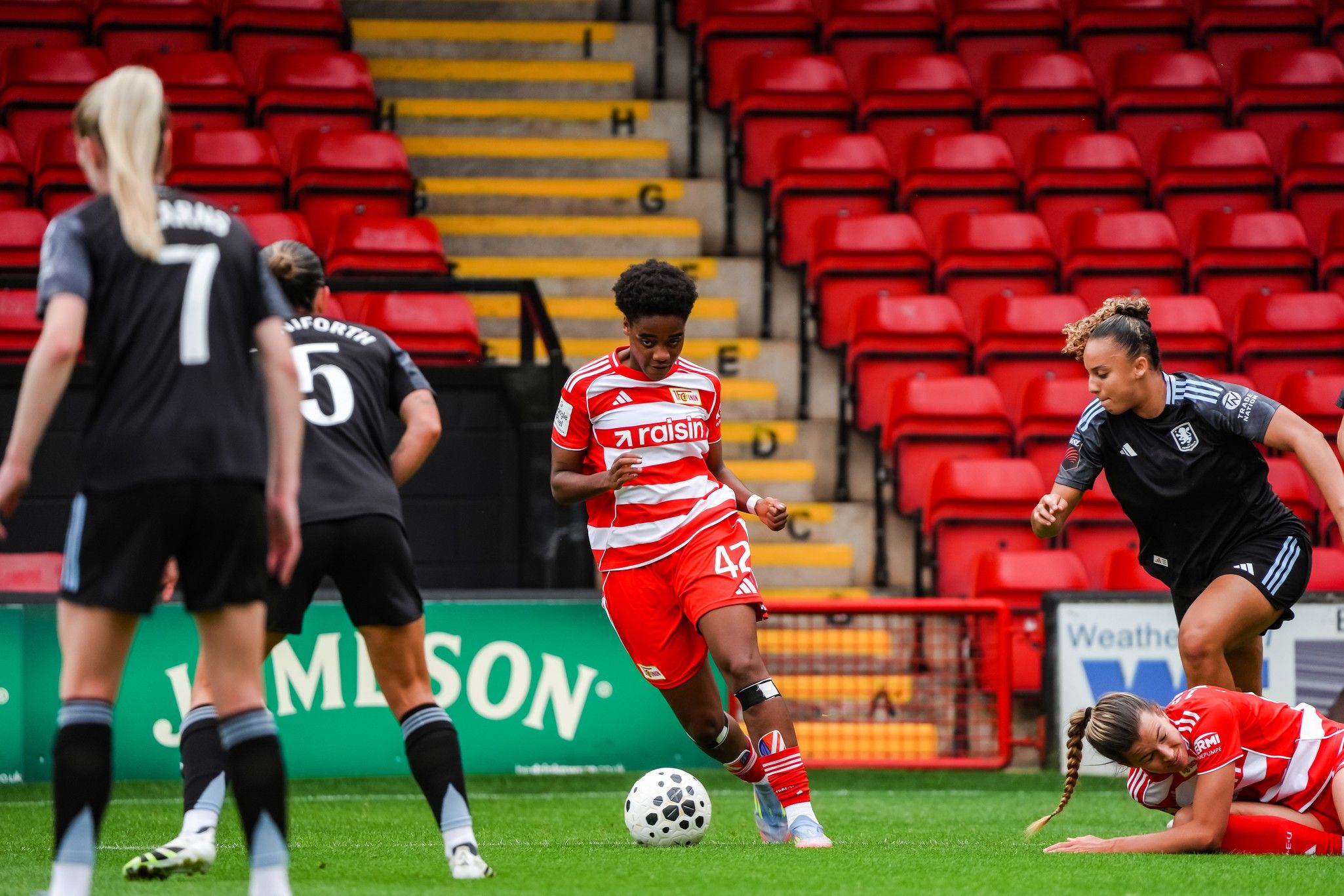 Eine Sportlerin in roter und weißer Kleidung dribbelt den Ball, während zwei Gegnerinnen versuchen, ihn zu erobern.