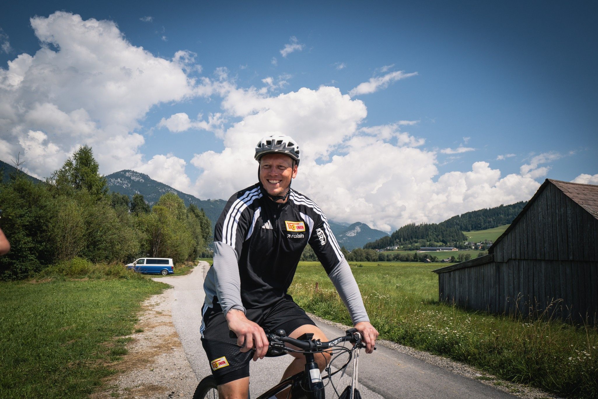 Ein Mann in Sportbekleidung und Helm sitzt auf einem Mountainbike, umgeben von einer ländlichen Landschaft mit Bergen und Wolken.