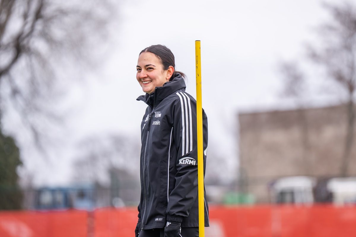 Lächelnde Sportlerin in schwarzer Trainingsjacke steht neben einer Trainingsstange auf einem Sportplatz im Freien.