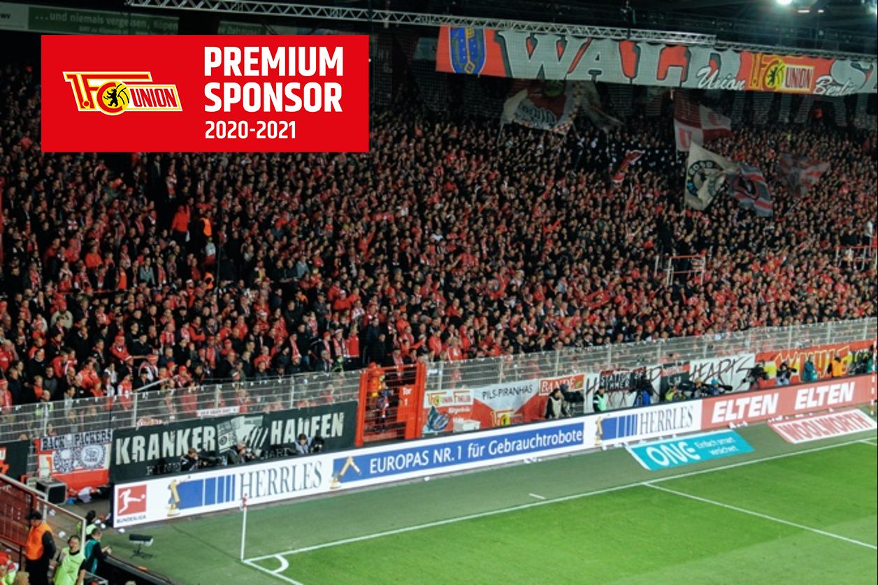 Ein vollbesetztes Stadion mit jubelnden Fans in roten Farben, Banner und Schildern im Hintergrund.