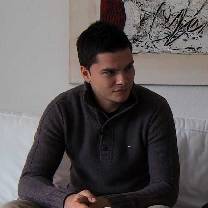 Ein junger Mann mit dunklen Haaren sitzt nachdenklich auf einer Couch vor einem kunstvoll gestalteten Wandbild.