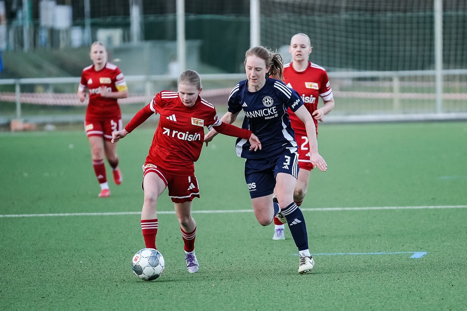 Zwei Fußballspielerinnen im Wettlauf um den Ball; eine in rotem und die andere in blauem Trikot. Hintergrund mit weiteren Spielerinnen.