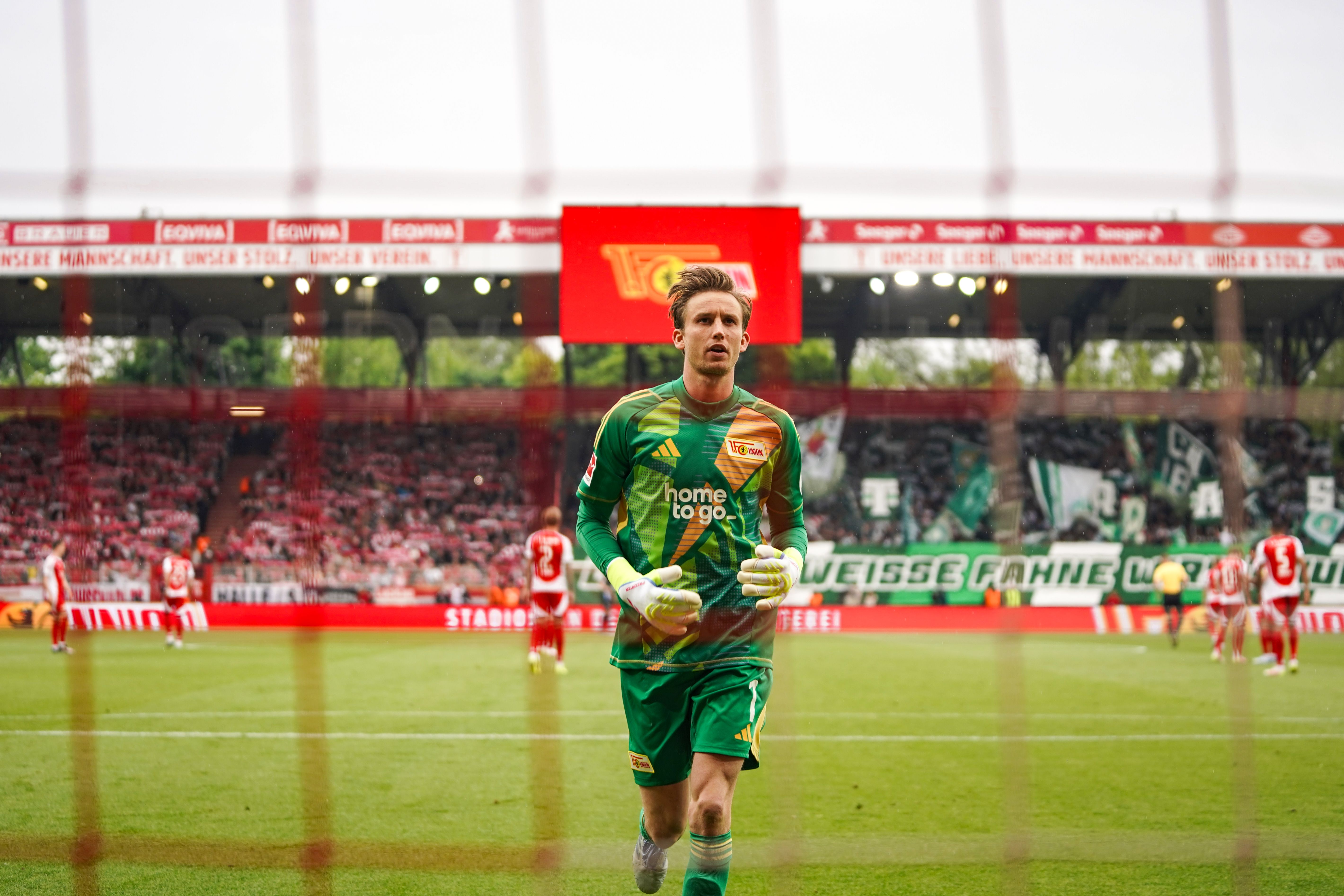 El portero con uniforme verde corre en el estadio, detrás de él los jugadores y aficionados en un partido de fútbol.