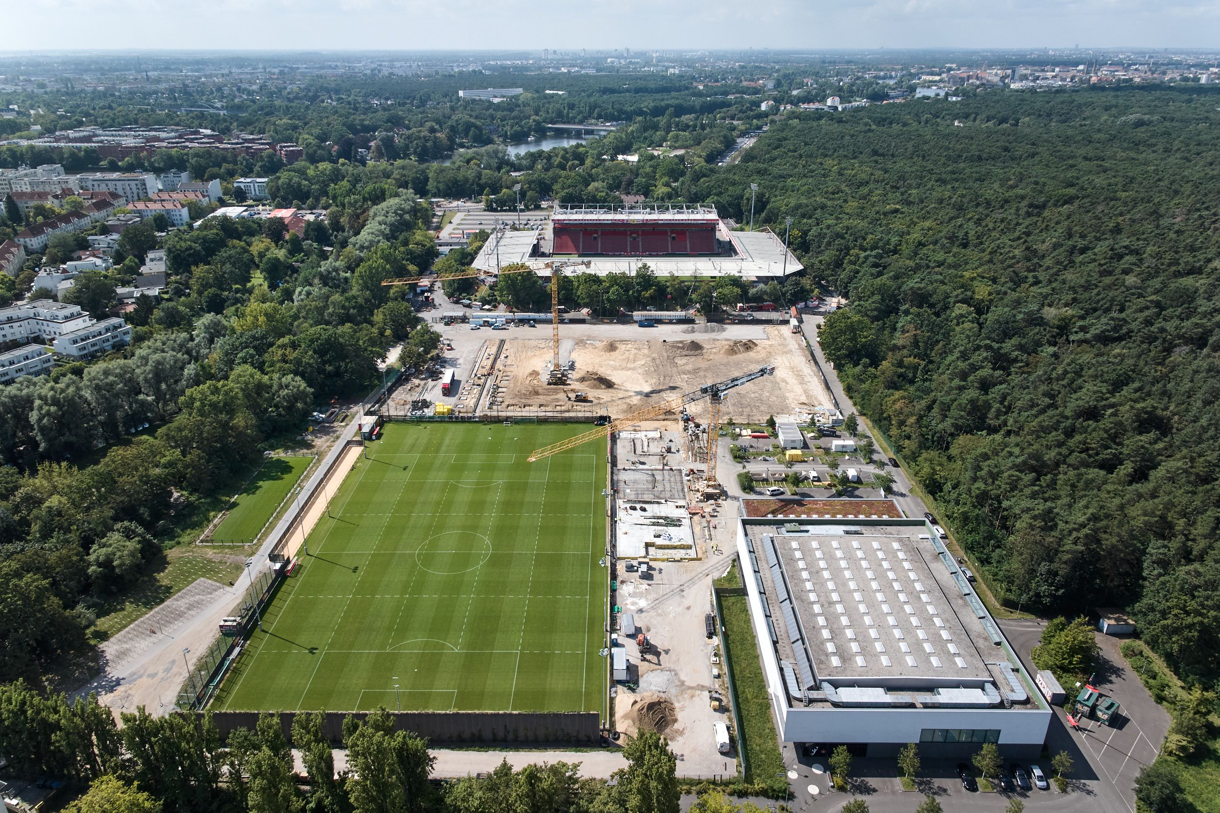 Aerial view of a sports facility with football fields, a stadium, and construction sites in an urban area with lots of greenery.