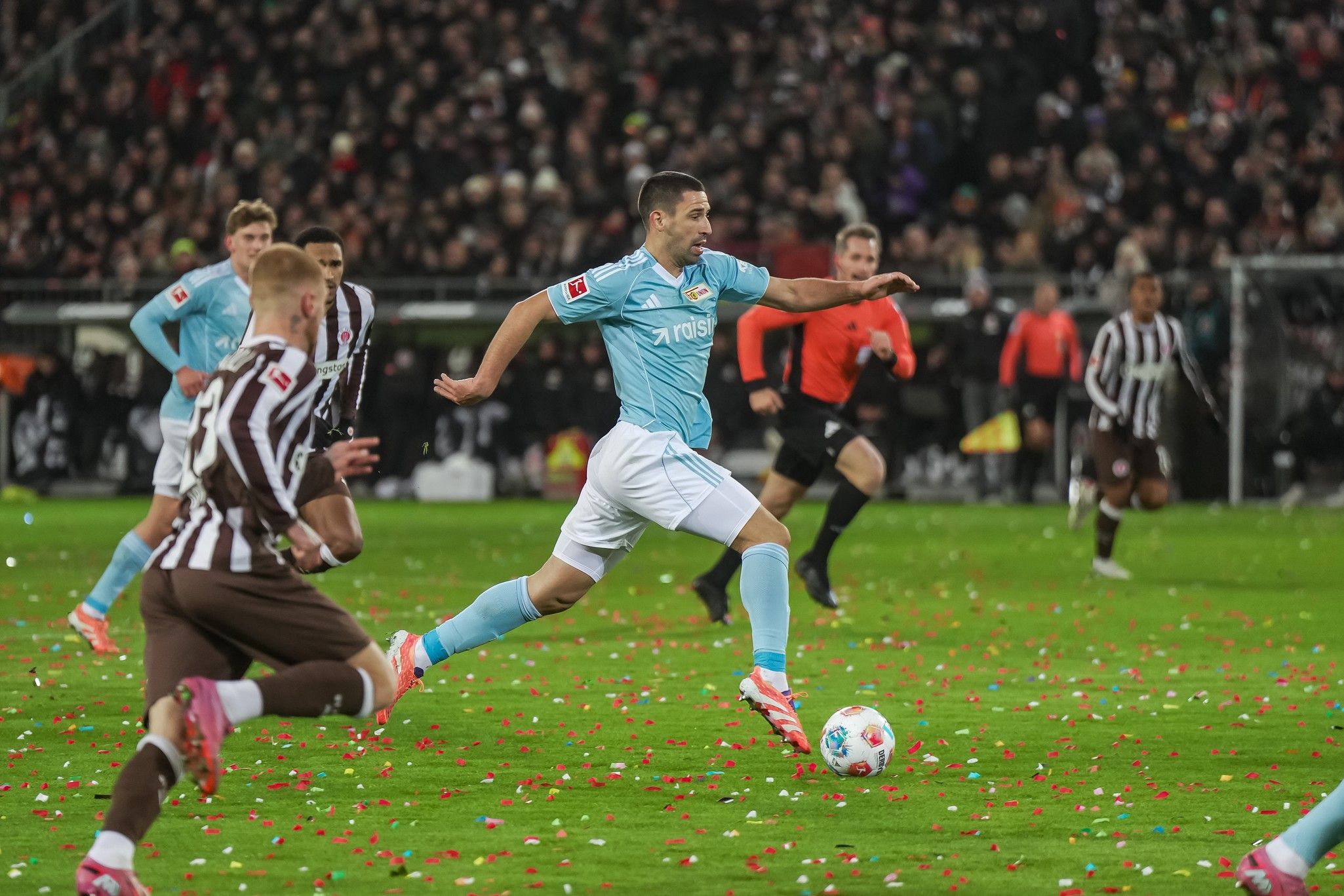 Spielszene im Fußball mit einem Spieler in hellblauem Trikot, der über das Feld sprintet, während bunte Konfetti auf dem Rasen liegen.