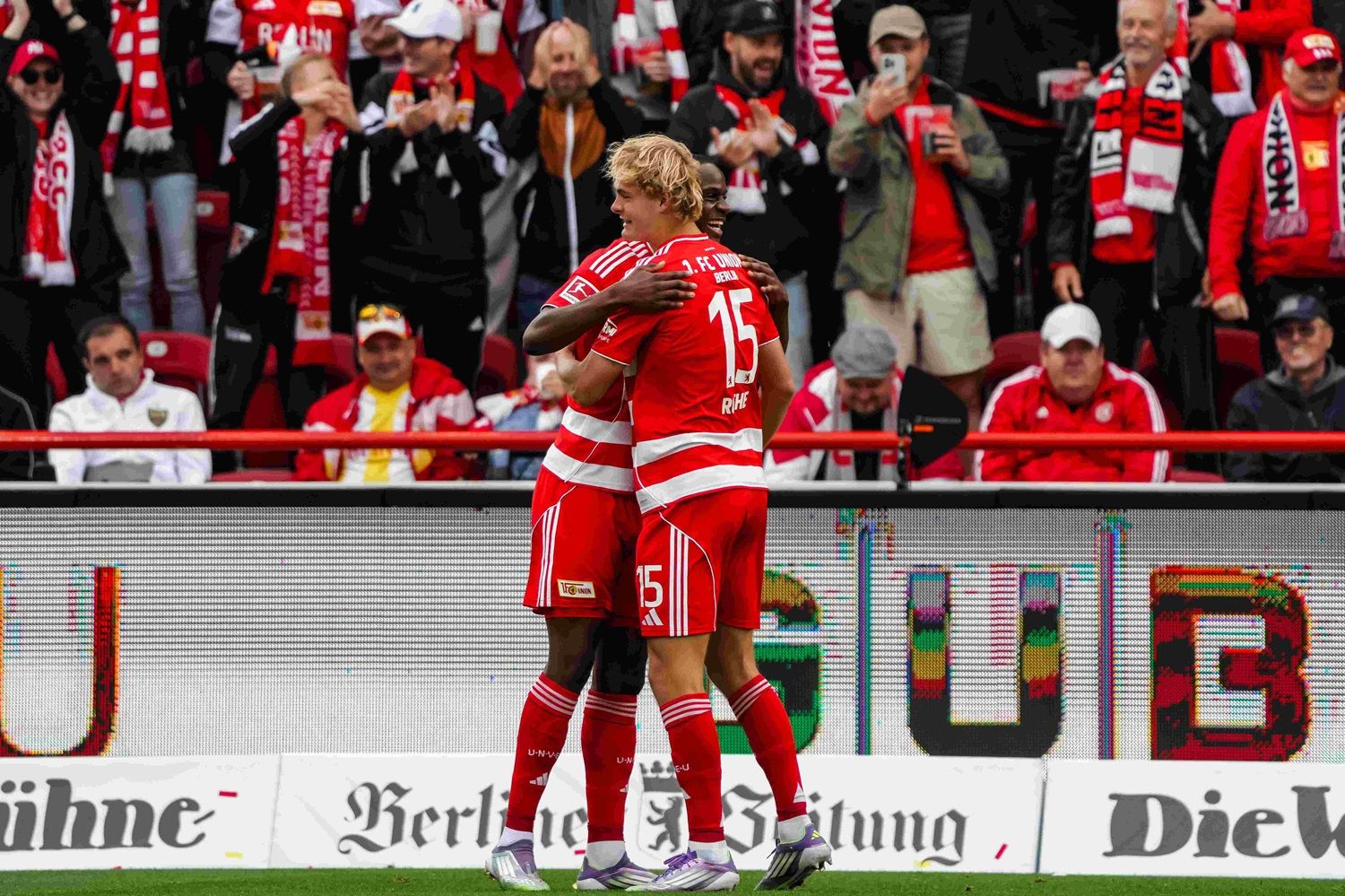 Zwei Fußballspieler umarmen sich jubelnd auf dem Spielfeld, während Fans im Hintergrund feiern.