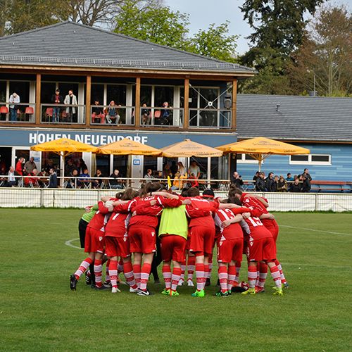 Spieler in roten Trikots bilden einen Kreis auf einem Fußballplatz, während Zuschauer in einem Gebäude und unter Sonnenschirmen zuschauen.