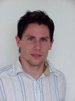 Ein junger Mann mit dunklen Haaren, der ein gestreiftes Hemd trägt, schaut direkt in die Kamera vor einem hellen Hintergrund.