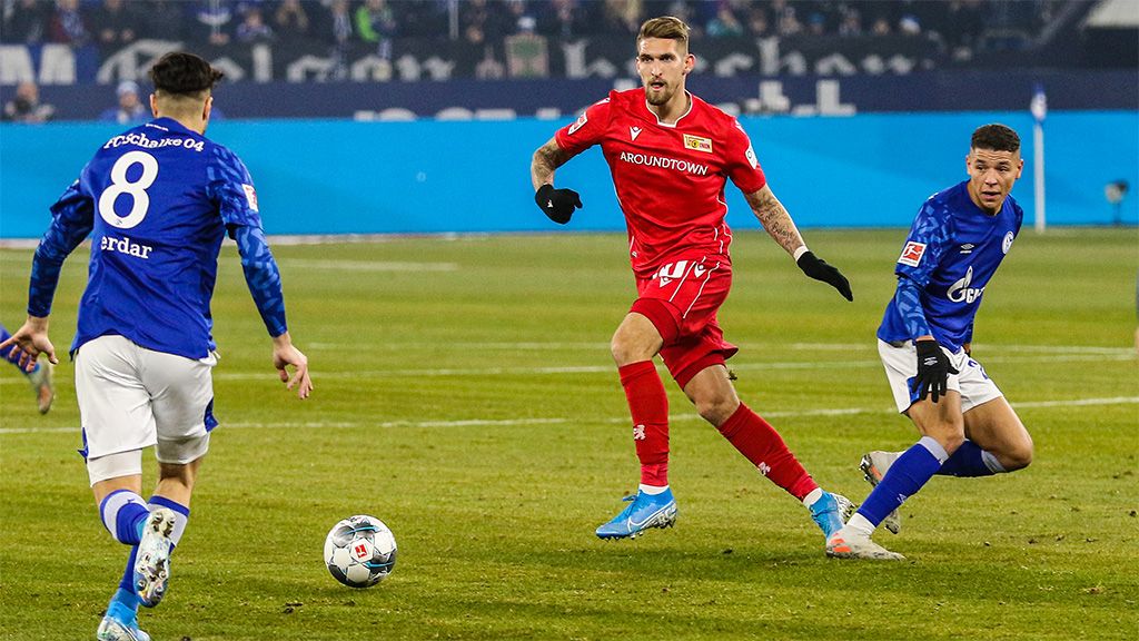 Zwei Fußballspieler in blauen Trikots verfolgen einen Spieler in rotem Trikot auf dem Feld während eines Spiels.