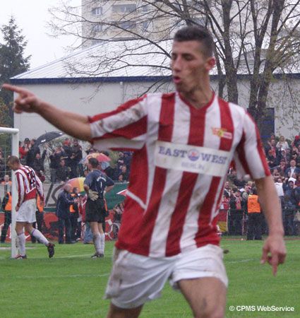 Ein Fußballspieler in rot-weiß gestreiftem Trikot jubelt und zeigt mit dem Finger auf einen Punkt. Im Hintergrund sind Zuschauer zu sehen.