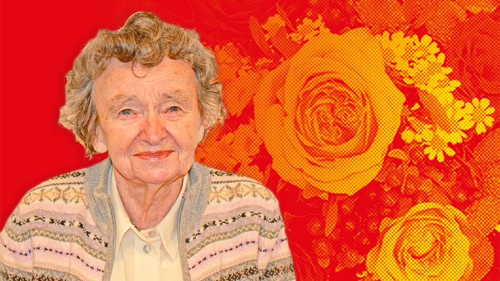 Eine ältere Frau mit lockigem, grauem Haar lächelt vor einem Hintergrund mit gelben Rosen und einem roten Farbverlauf.