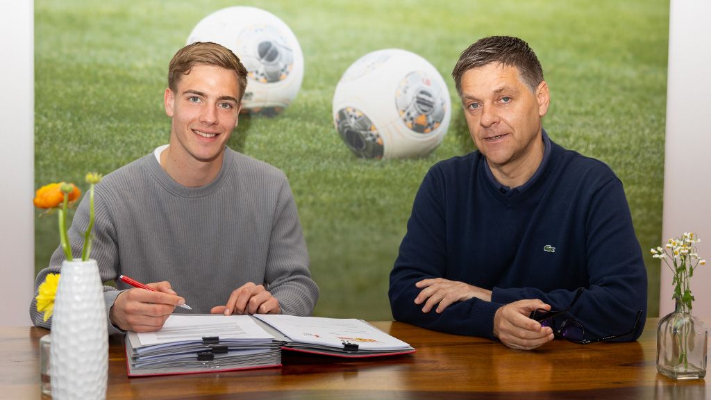 Two men are sitting at a table, one is signing documents, and in the background, football and grass can be seen.
