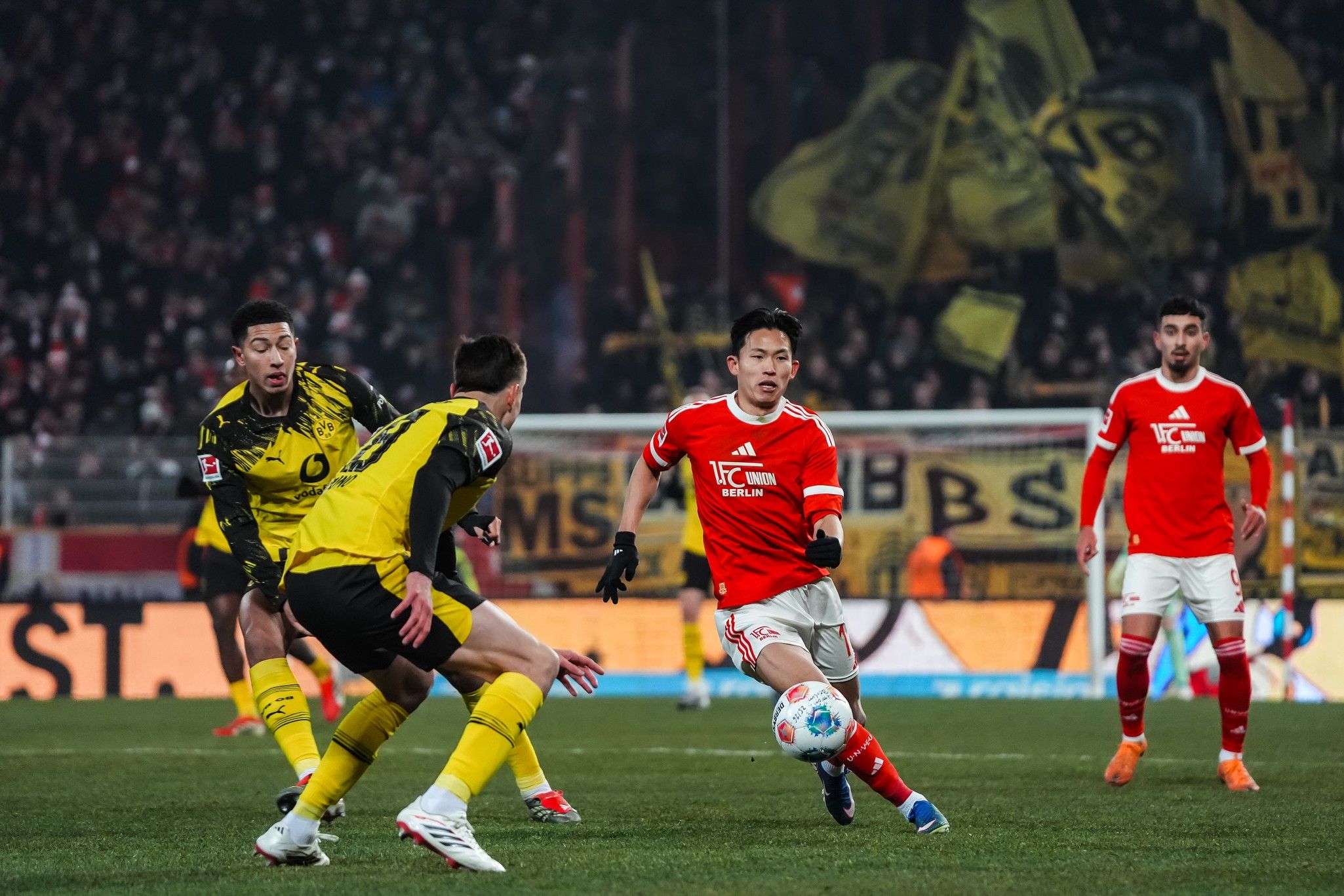 Fußballspiel zwischen zwei Mannschaften, in dem ein Spieler den Ball führt, während andere Spieler in Aktion sind. Hintergrund mit Fans.