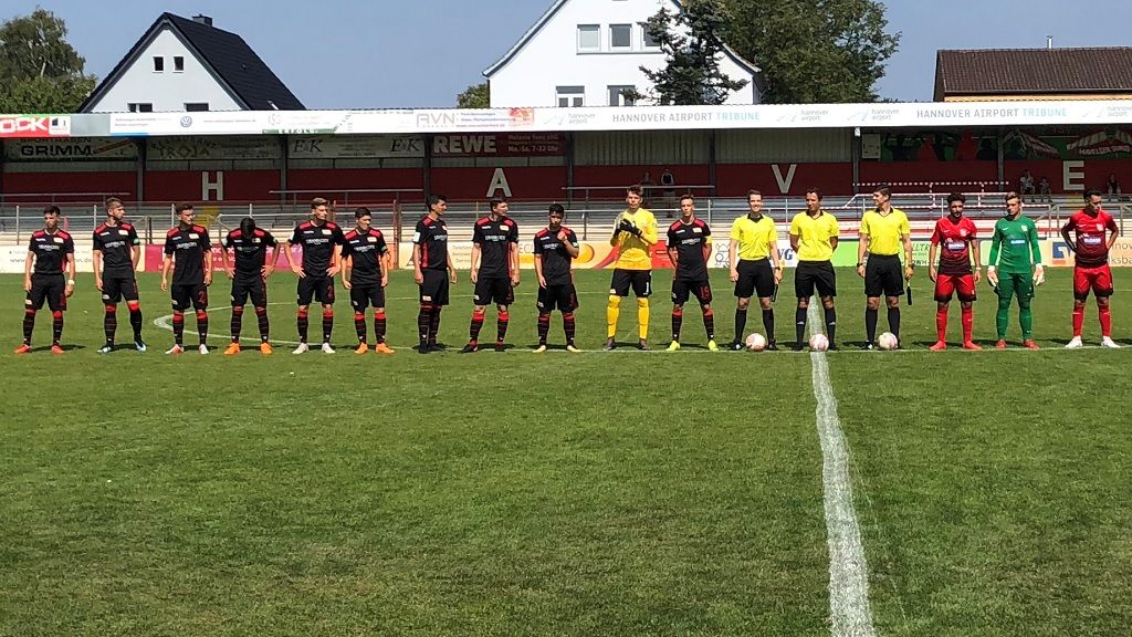 Zwei Fußballmannschaften stehen auf dem Feld in Reihen aufgestellt, bereit zum Spiel. Schiedsrichter in der Mitte.