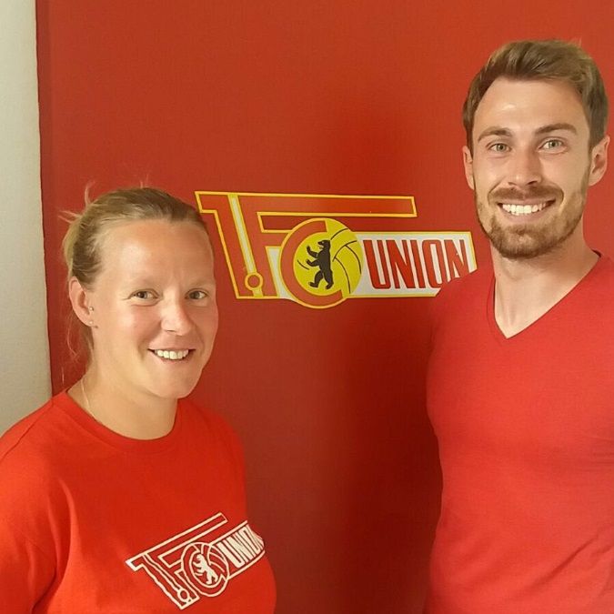 Zwei Personen in roten T-Shirts stehen vor einer rot-gelben Wand mit dem Logo von "1. FC Union".