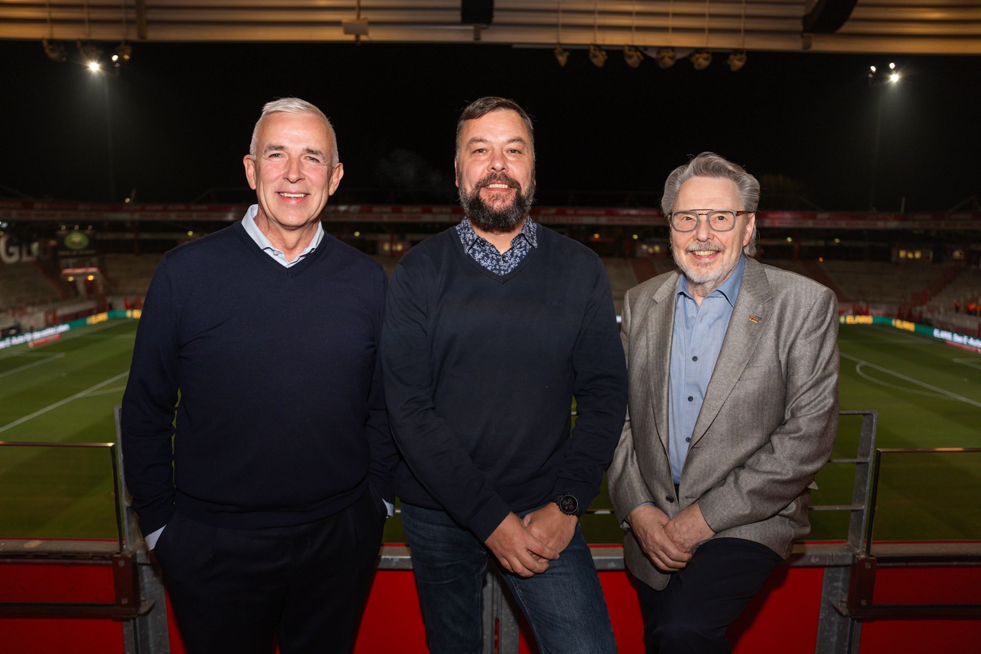 Drei Männer stehen in einem Stadion mit beleuchtetem Spielfeld im Hintergrund, lächeln und posieren für das Foto.