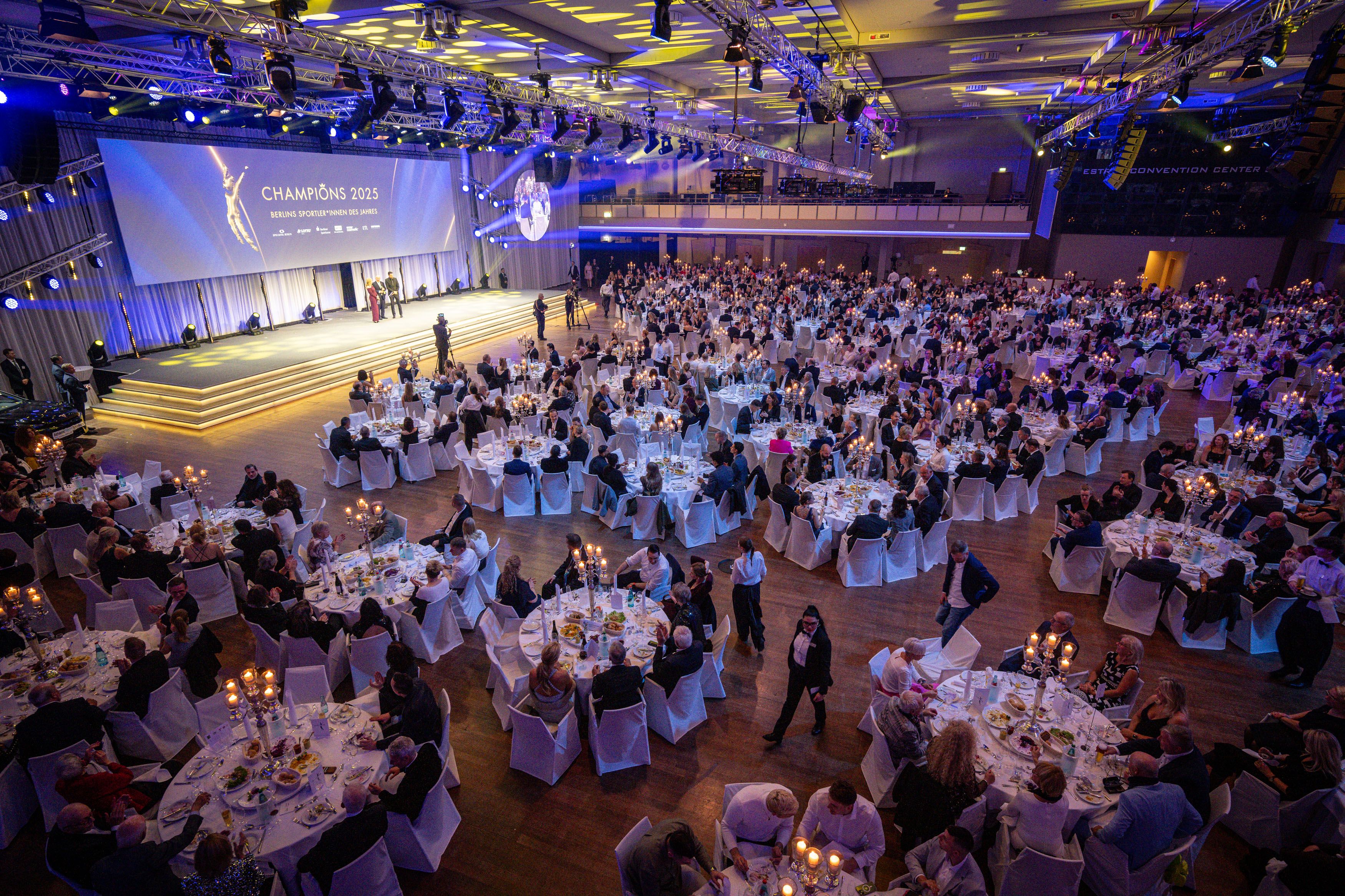 Eine große Gala mit zahlreichen Gästen, festlich gedeckten Tischen und einer Bühne im Hintergrund.