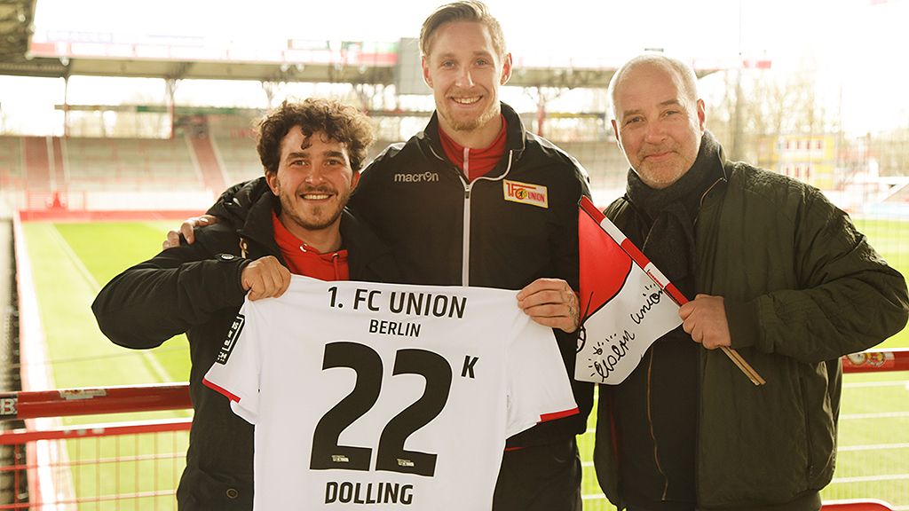 Drei Männer halten ein Trikot des 1. FC Union Berlin mit der Nummer 22 und dem Namen Dolling in einem Stadion.