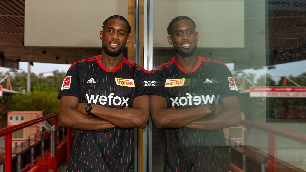 Ein Mann steht mit verschränkten Armen vor einer Glasscheibe, reflektiert in der Scheibe. Er trägt ein schwarzes Fußballtrikot.