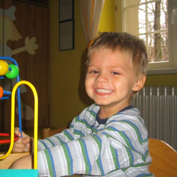 Ein lächelnder Junge sitzt an einem Tisch und spielt mit einem bunten Holzspielzeug. Hintergrund mit hellen Wänden.