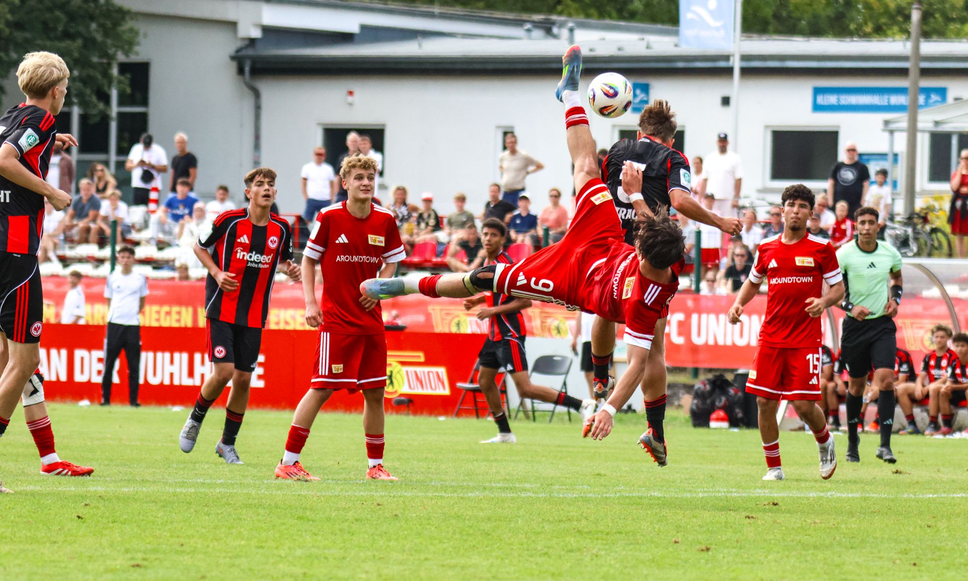Spielszene im Fußball mit einem Spieler, der einen spektakulären Rückwärtssalto schießt, während andere Spieler zuschauen.