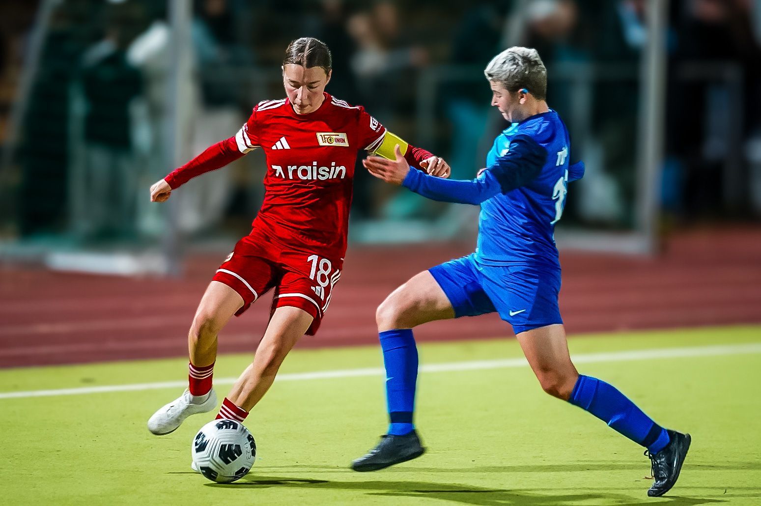 Spielszene im Fußball: Eine Spielerin in Rot dribbelt den Ball, während eine Spielerin in Blau eingeholt wird.