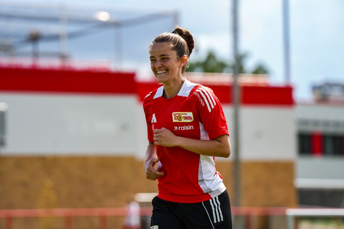 Eine lächelnde Fußballspielerin trägt ein rotes Trikot und läuft auf einem Sportplatz mit Trainingsausrüstung im Hintergrund.