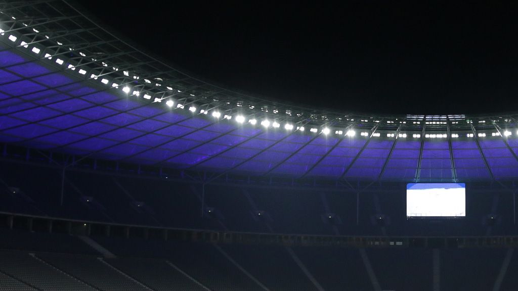 Ein beleuchtetes Stadiondach bei Nacht mit einem großen Bildschirm und leeren Sitzplätzen.