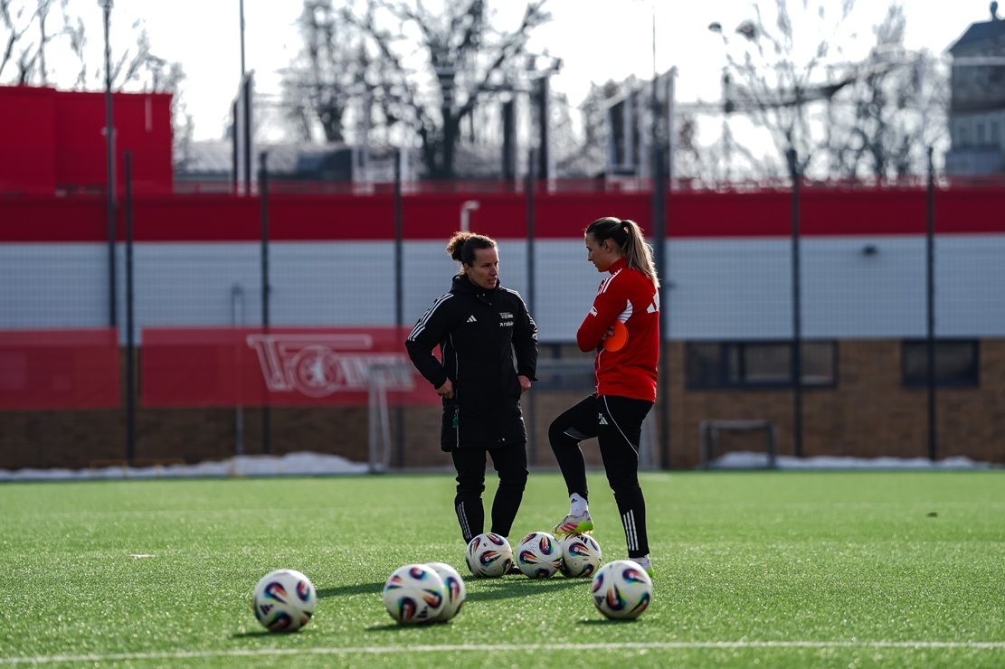 Zwei Fußballspielerinnen im Training stehen auf einem Feld, umgeben von mehreren Fußbällen.