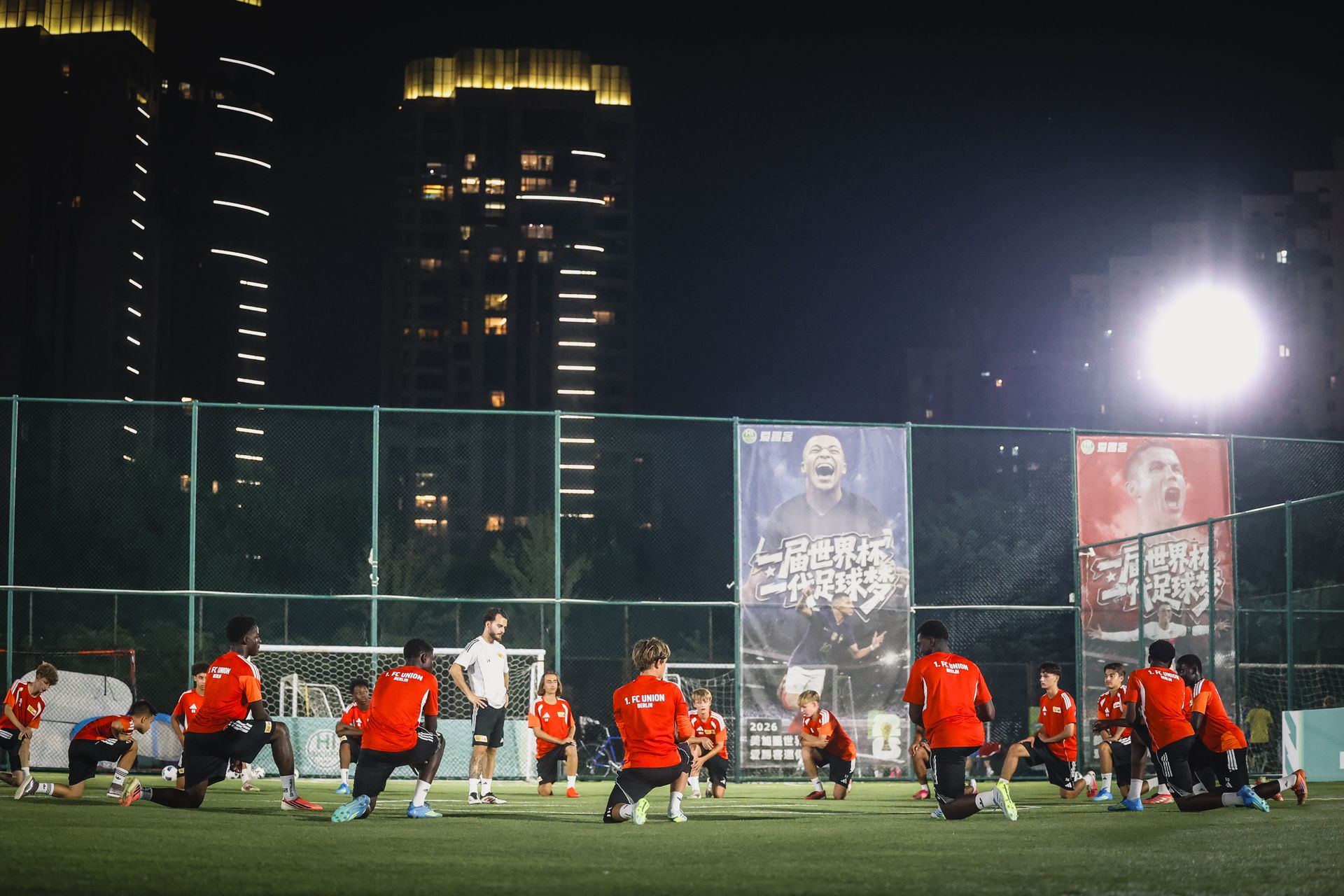 Fußballmannschaft in roten Trikots trainiert bei Nacht auf einem Platz mit Stadtlichtern im Hintergrund.