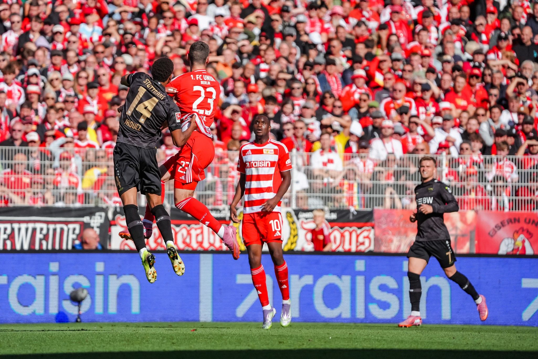 Spielszene im Fußballstadion: Spieler springen um den Ball, im Hintergrund eine jubelnde Menschenmenge.